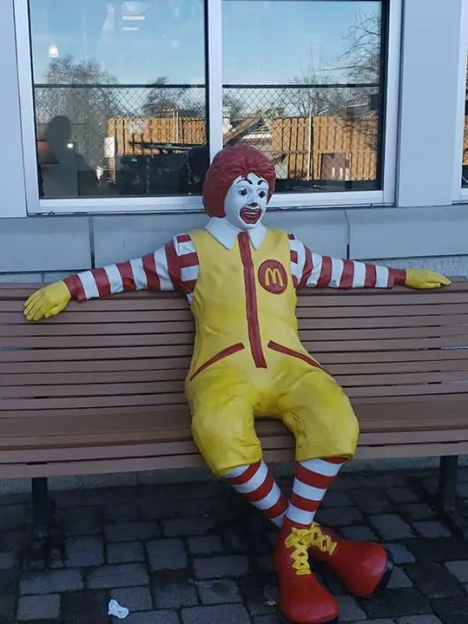 A statue of Ronald McDonald, a clown with red hair and a yellow outfit, sitting on a bench with his arms resting on the back of the bench.
