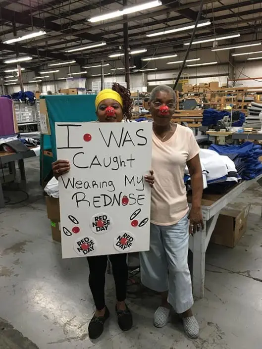 Two women wearing red noses. One of them is holding a sign that says "I was caught wearing my red nose".