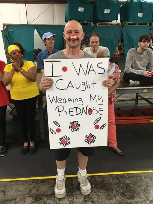 A man wearing a red nose. He is holding a sign that says "I was caught wearing my red nose".
